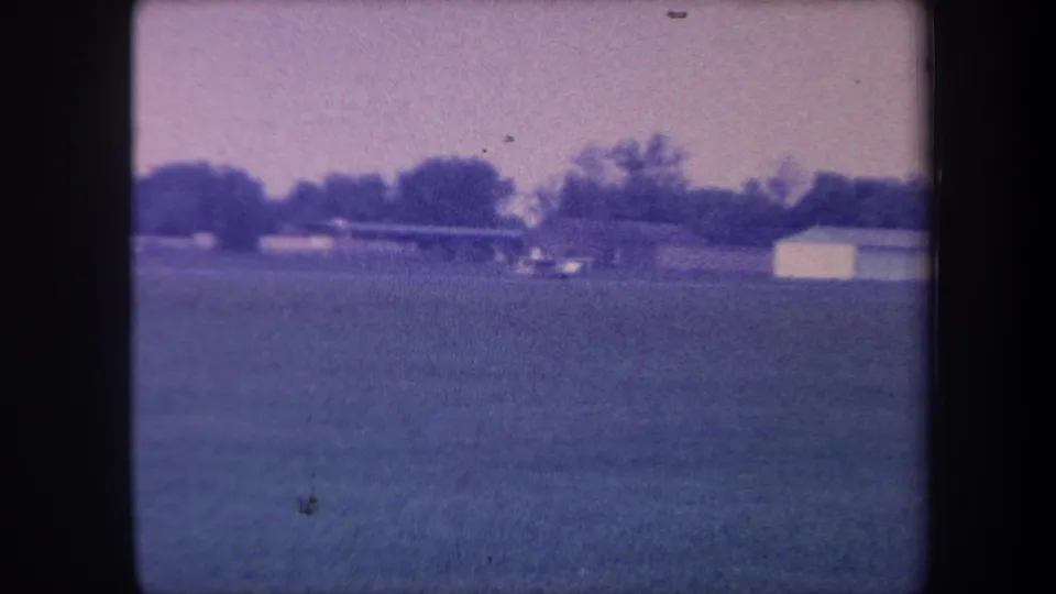 8mm home movie frame, circa 1959, Yucaipa, California area; small aircraft slowing near a white hangar structure at the far edge of the field.