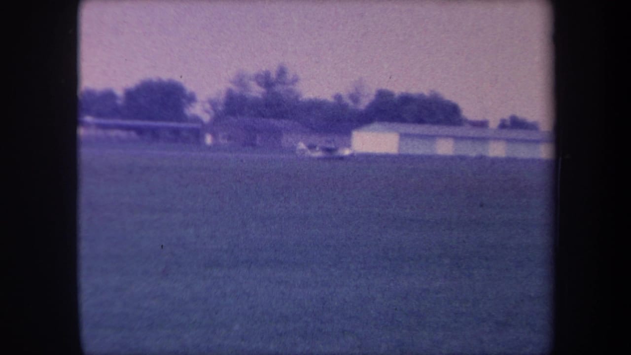 Static 8mm home movie frame from 1959 Yucaipa, California — open field, low outbuilding, and treeline under a hazy sky, with purple-blue emulsion shift.