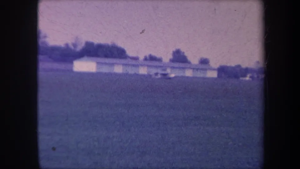 Continuation frame from 1959 Yucaipa, California — open field with low outbuilding and treeline; a small vehicle or machine appears in the middle distance.