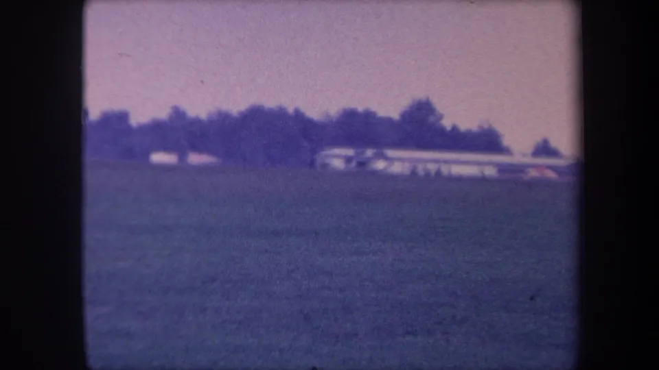 Small low-wing single-engine aircraft skimming low over a grass airfield, possibly near Yucaipa, California, circa 1959; strong purple dye-shift from aged 8mm film.