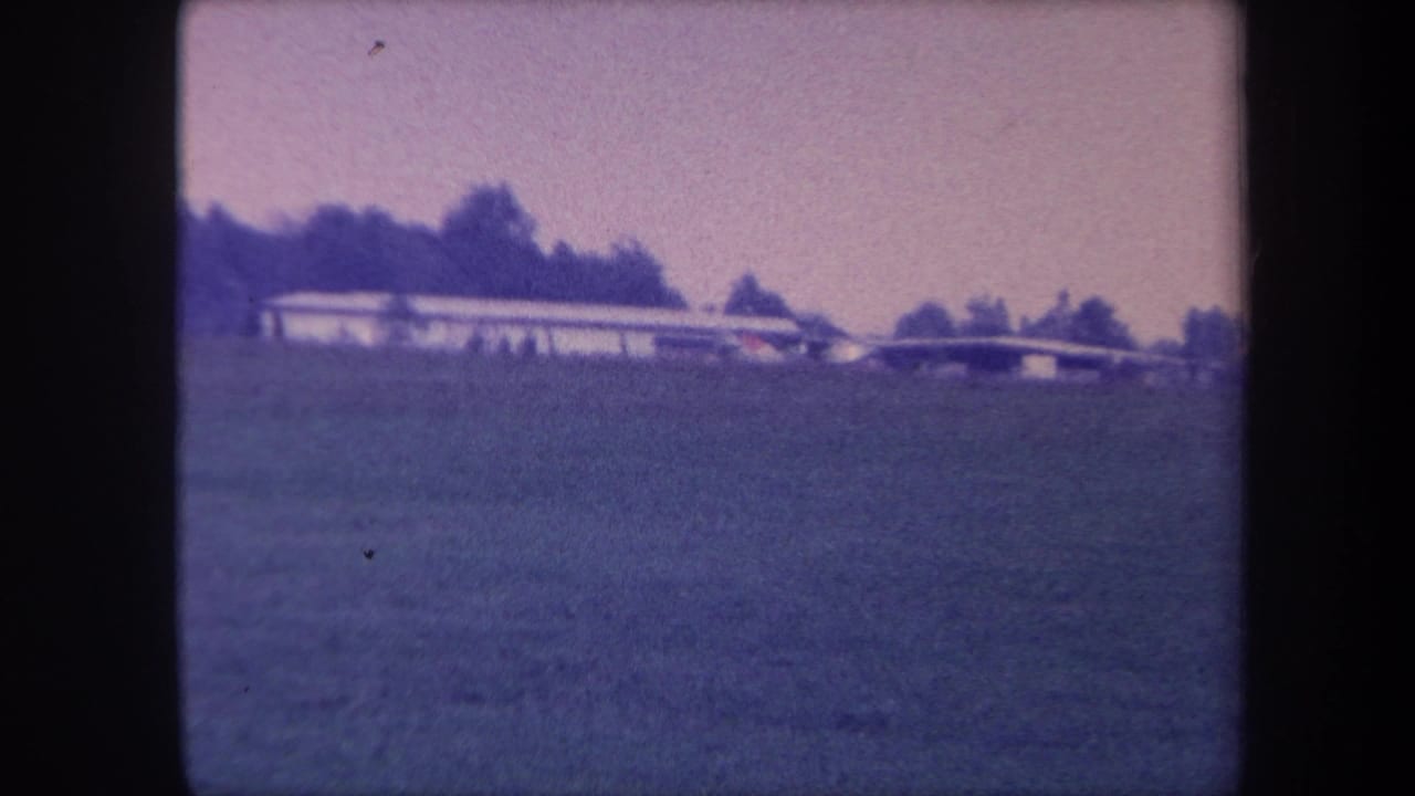Establishing shot of a grass airfield, possibly Yucaipa, California, circa 1959; long low building at left and tree-lined horizon on aged 8mm film with purple color cast.
