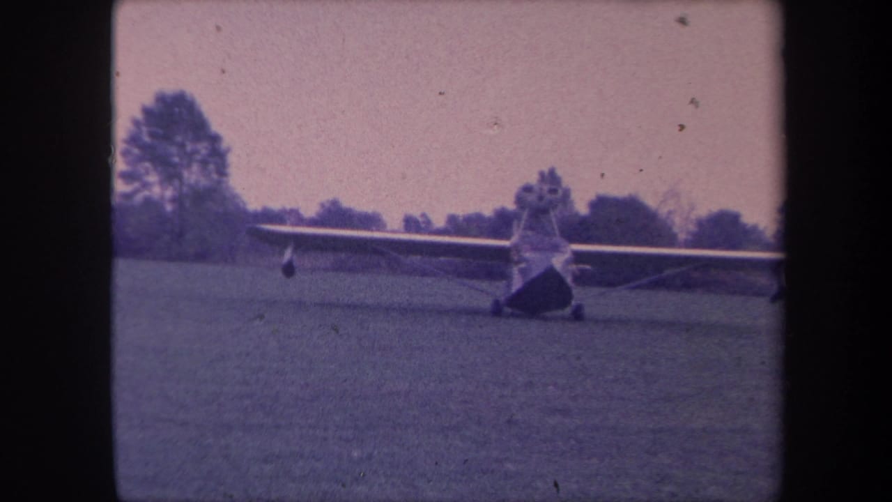 High-wing light aircraft on grassy airfield, slightly tighter framing than preceding frame, same blue-magenta aged 8mm color cast.