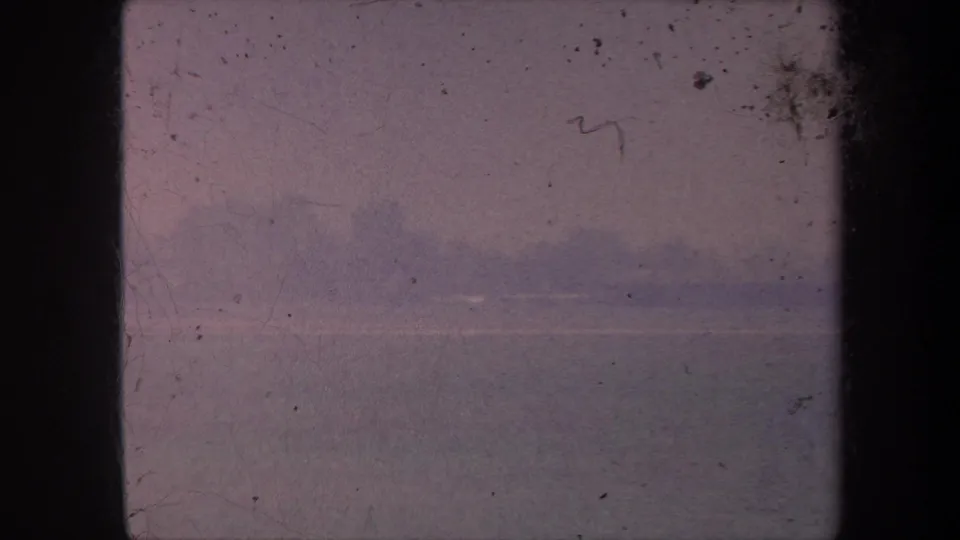 1959 8mm home movie frame: open horizon with pink-mauve sky; dust spots and emulsion scratches concentrated in the upper-right corner.