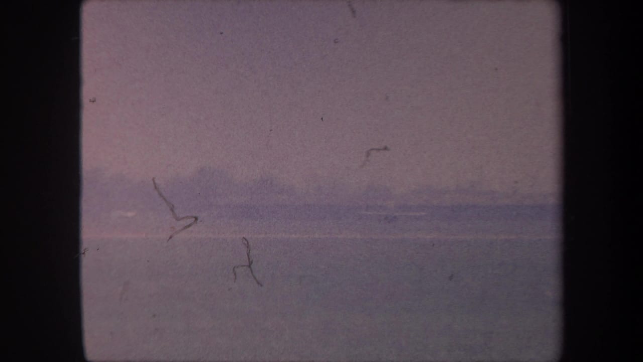 Heavily faded 8mm frame showing a purple-toned hazy sky above a flat horizon, with film scratches and indistinct marks at left.