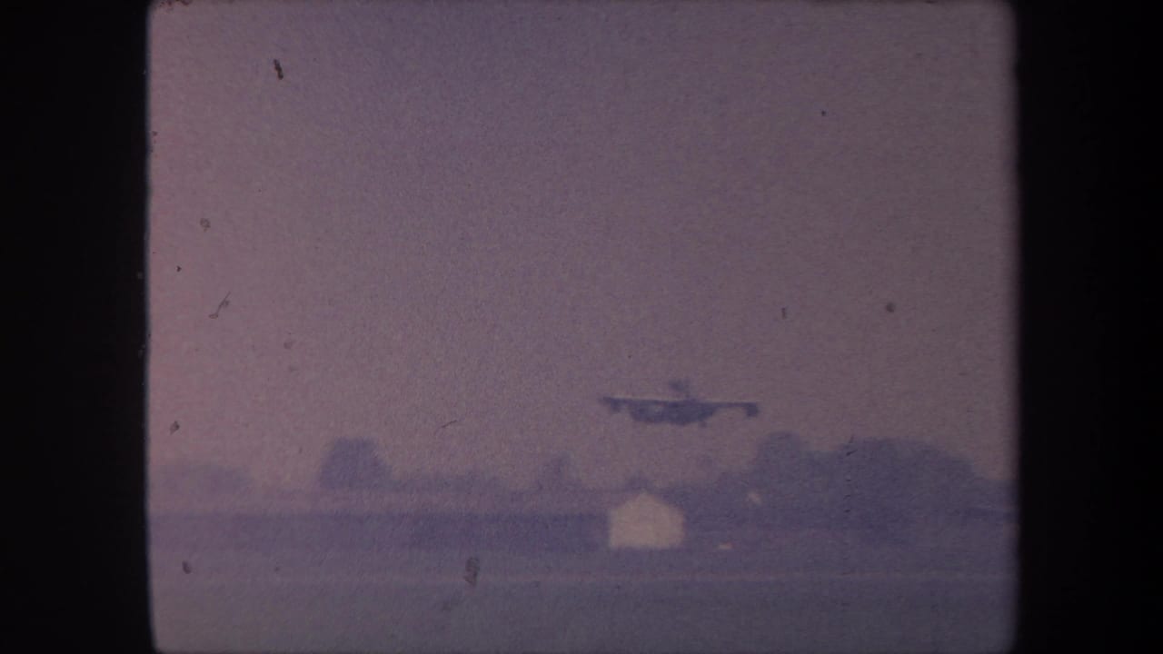 Small propeller aircraft just above ground level with a white building and treeline on the horizon, purple-tinted 8mm film.