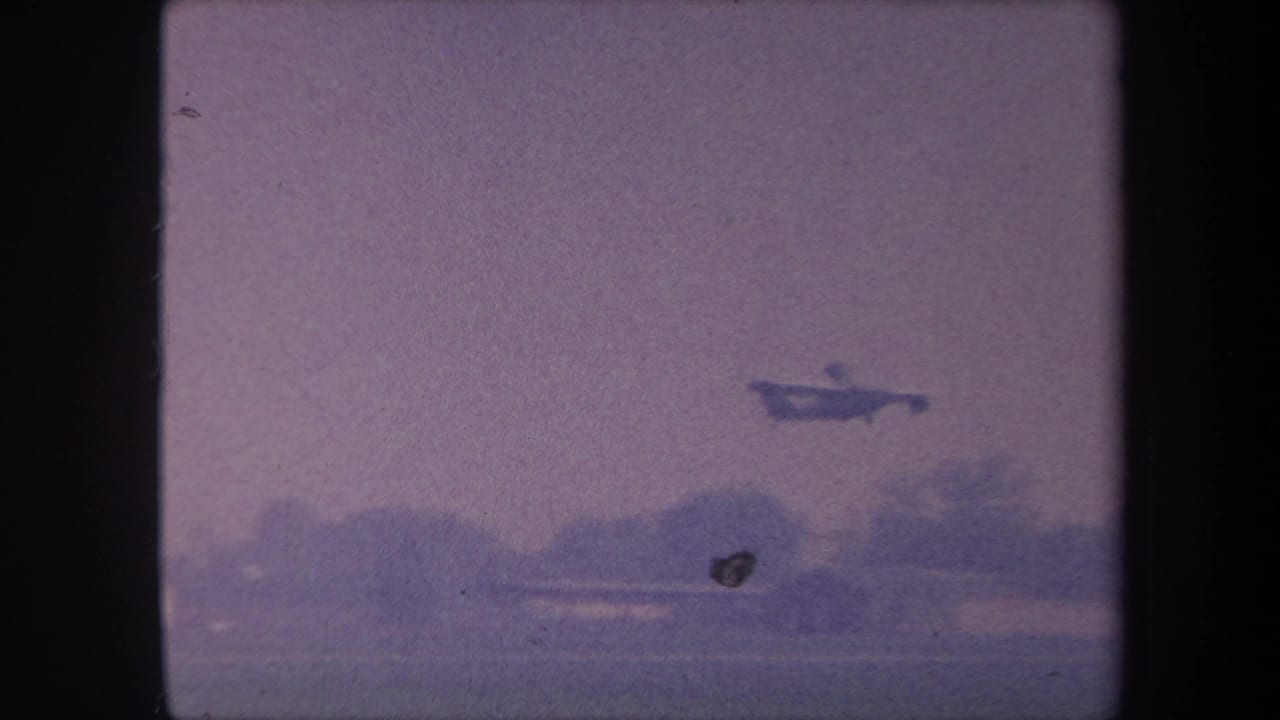 Small propeller aircraft in mid-climb over rural landscape circa 1959, with a second dark airborne object visible — possibly a bird or film artifact — shot on 8mm with heavy purple color degradation.