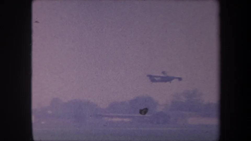 Small propeller aircraft in mid-climb over rural landscape circa 1959, with a second dark airborne object visible — possibly a bird or film artifact — shot on 8mm with heavy purple color degradation.