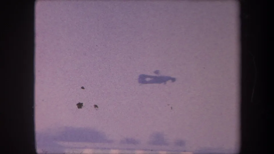 Small propeller aircraft in flight against a hazy lavender sky, filmed on 8mm circa 1959 with scattered film debris specks and color degradation evident.