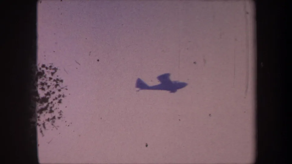 Small propeller aircraft fully airborne against a lavender sky circa 1959, with a heavy emulsion debris cluster at frame left indicating significant film-stock deterioration.