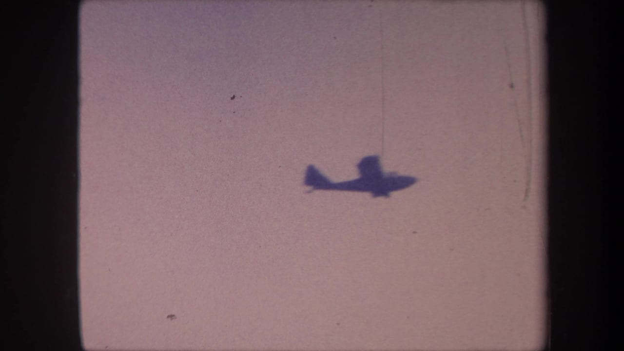 Small propeller aircraft silhouette in upper-left of pale overcast sky, 8mm film-frame borders and dust specks visible.