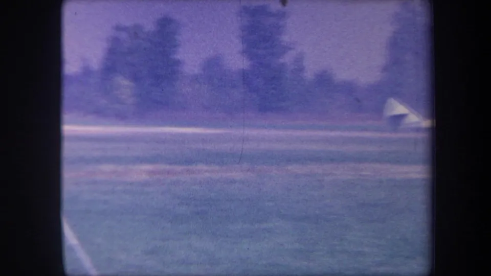 Wide grass airstrip view with aircraft barely visible at distance; a curved foreground element appears at the lower edge of this circa-1959 8mm frame.