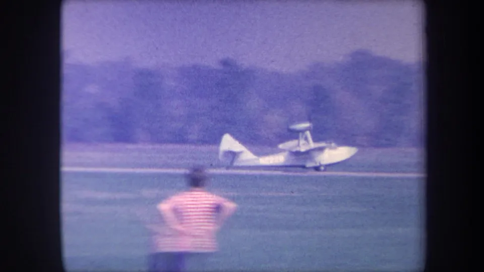 An unidentified adult seen from behind in a striped shirt watches a seaplane on a grass strip, likely Yucaipa, California, circa 1959; distant hills visible through haze.