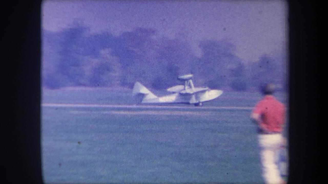 Small white amphibious aircraft taxiing on a grass airfield, metadata-identified as Yucaipa, California, circa 1959; one bystander in red shirt visible from behind.