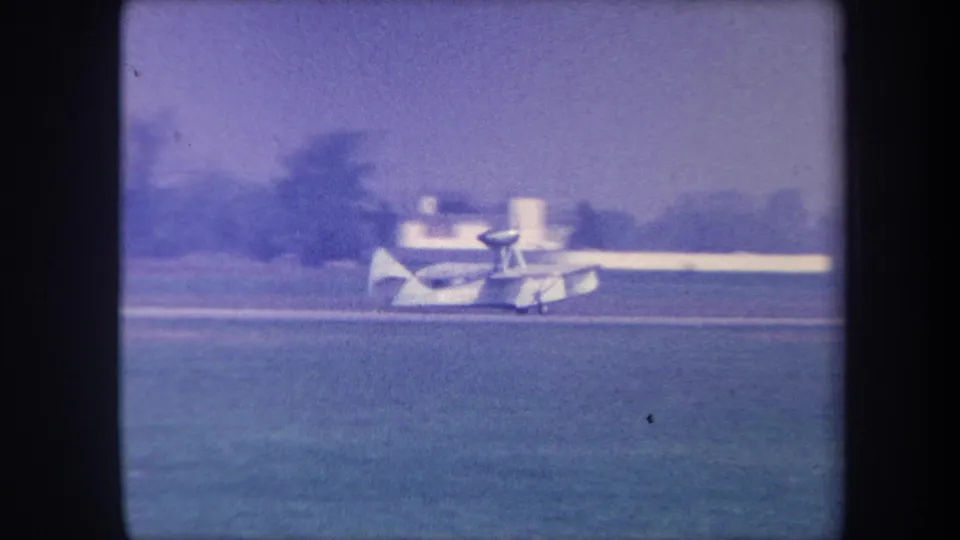 Small white amphibious aircraft taxiing across a grass airfield, circa 1959; the bystander from the opening frame has moved out of view.