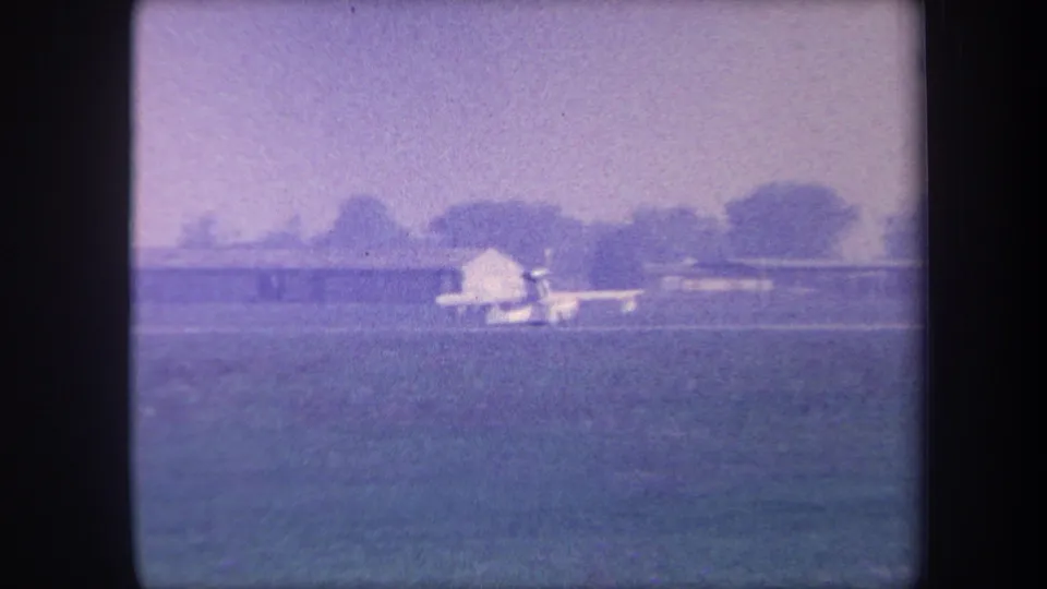 Light aircraft on rural grass airstrip with a low white outbuilding behind it and tree line at horizon, 8mm home movie circa 1959, possibly Yucaipa, California.