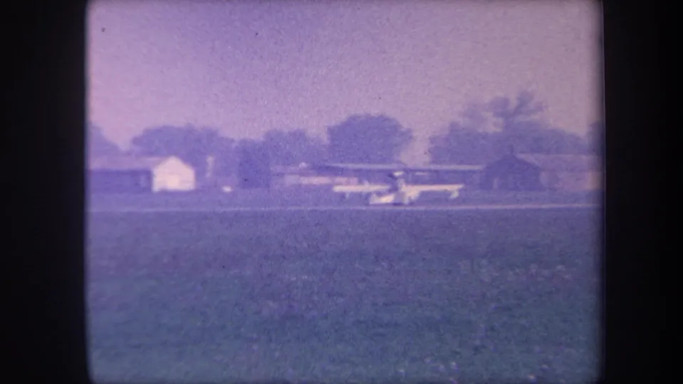Light aircraft at or near liftoff on a rural grass strip with a low white building at right and tree line behind, 8mm home movie circa 1959, possibly Yucaipa, California.
