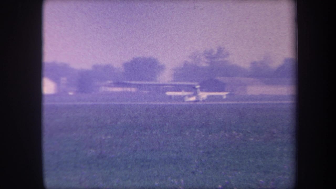 Small single-engine plane on a grassy airstrip beside a low flat-roofed hangar, trees in background, heavy purple film cast.