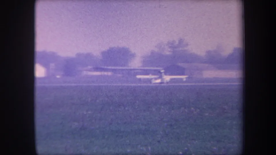Small propeller aircraft on a flat grassy airstrip near a low hangar, Yucaipa California area, circa 1959, 8mm home movie with pronounced purple color shift.