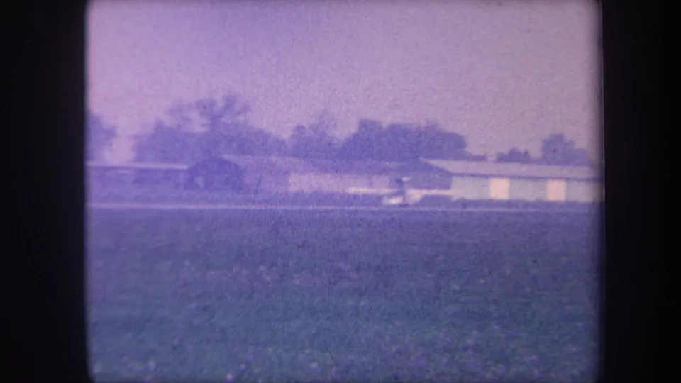 Wide shot of a flat grassy airstrip with a low-slung hangar structure and tree line to the rear, Yucaipa area California, circa 1959, 8mm home movie footage.
