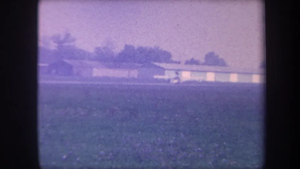 Low-slung hangar building at the edge of a flat grassy airstrip, tree-lined background, Southern California area, circa 1959, aged 8mm home movie footage.