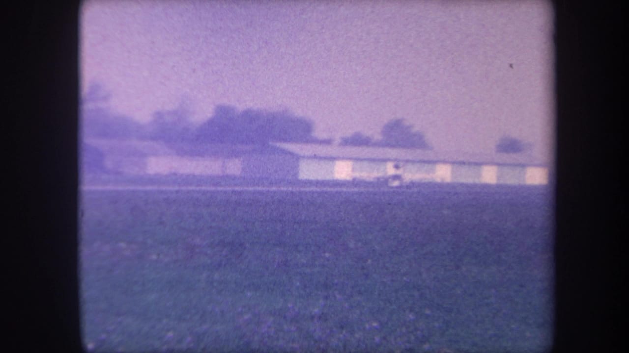 Continuation of a 1959 8mm sequence at a rural California airfield; hangar and field composition nearly unchanged from the opening frame.