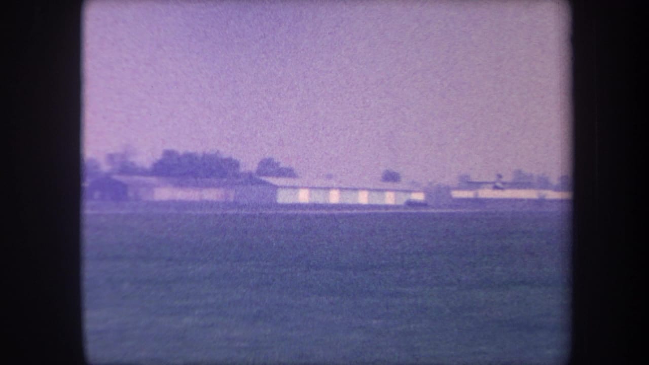 Wide view of a rural California airfield showing low elongated hangars and a sparse treeline, captured on 8mm color film circa 1959 with strong purple dye-fading.