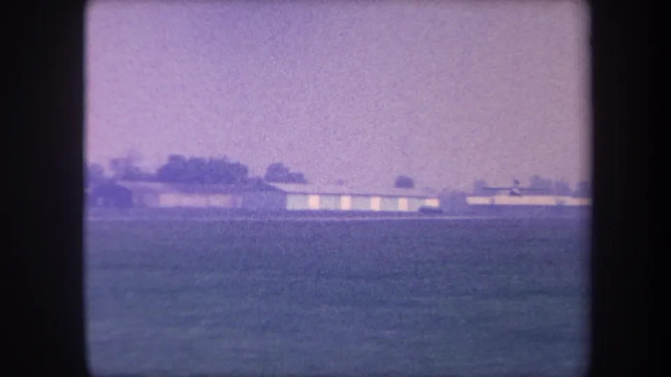 Wide view of a rural California airfield showing low elongated hangars and a sparse treeline, captured on 8mm color film circa 1959 with strong purple dye-fading.