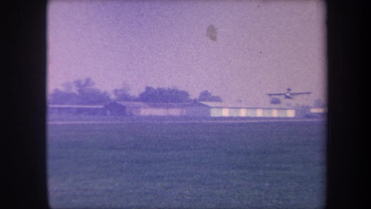 Small propeller aircraft at the moment of liftoff from a rural airfield in the Yucaipa area, California, circa 1959, with hangars and treeline visible on 8mm home movie film.