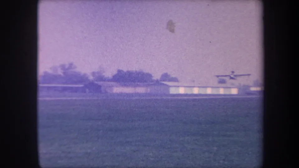 Small propeller aircraft at the moment of liftoff from a rural airfield in the Yucaipa area, California, circa 1959, with hangars and treeline visible on 8mm home movie film.