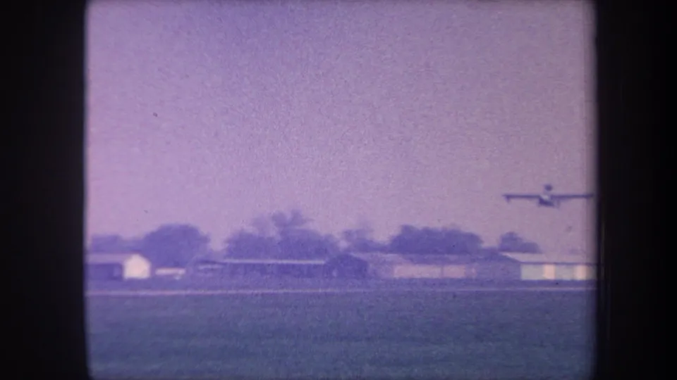 Light propeller aircraft climbing slowly above the grass strip and hangars of a rural California airfield, circa 1959, captured on aged 8mm home movie film.