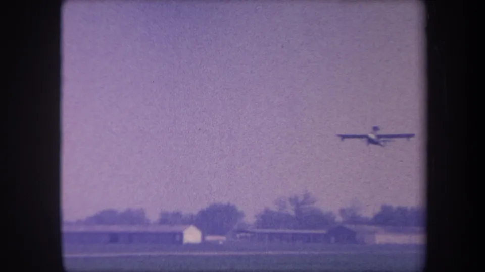 Light aircraft continuing its slow climb and receding over the rural Yucaipa-area airfield, with hangars and treeline below, on 8mm home movie film circa 1959.