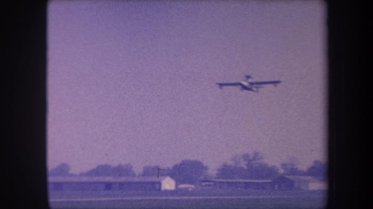 Single-engine propeller plane flying left-to-right in a purple-tinted sky above a flat rural landscape with low buildings and trees at the horizon.
