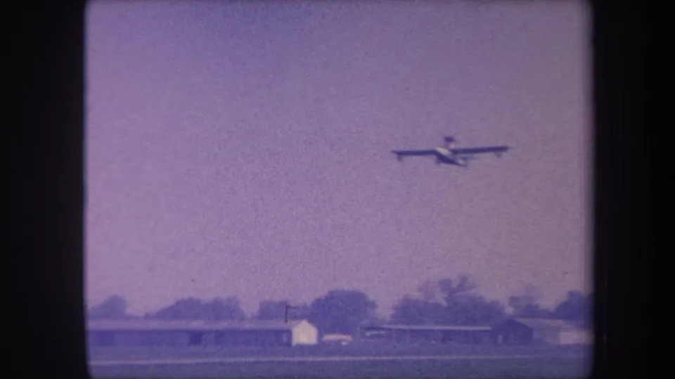 Light propeller aircraft in flight over a rural California airfield, 1959 8mm home movie, purple dye-shifted sky with low hangar building and treeline below.