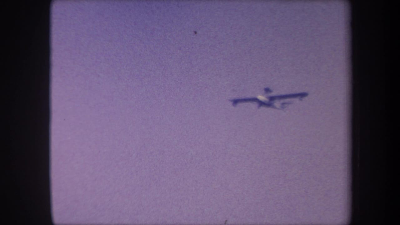 Light propeller aircraft mid-flight, 1959 8mm home movie, sky-fill frame with purple dye shift and no horizon visible.