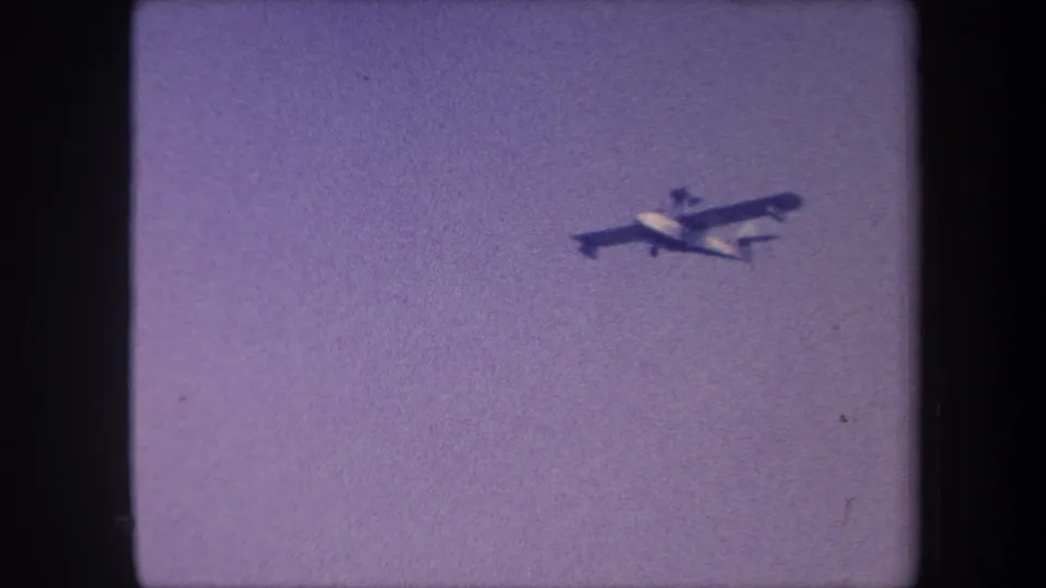 Light aircraft in closer view, 1959 8mm home movie, purple-tinted sky, high-wing monoplane profile more distinct as camera tracks subject.