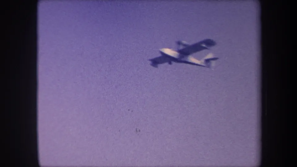 Light prop plane at closest framing in the clip, 1959 8mm footage, California, underside and wing profile visible against purple-cast sky.