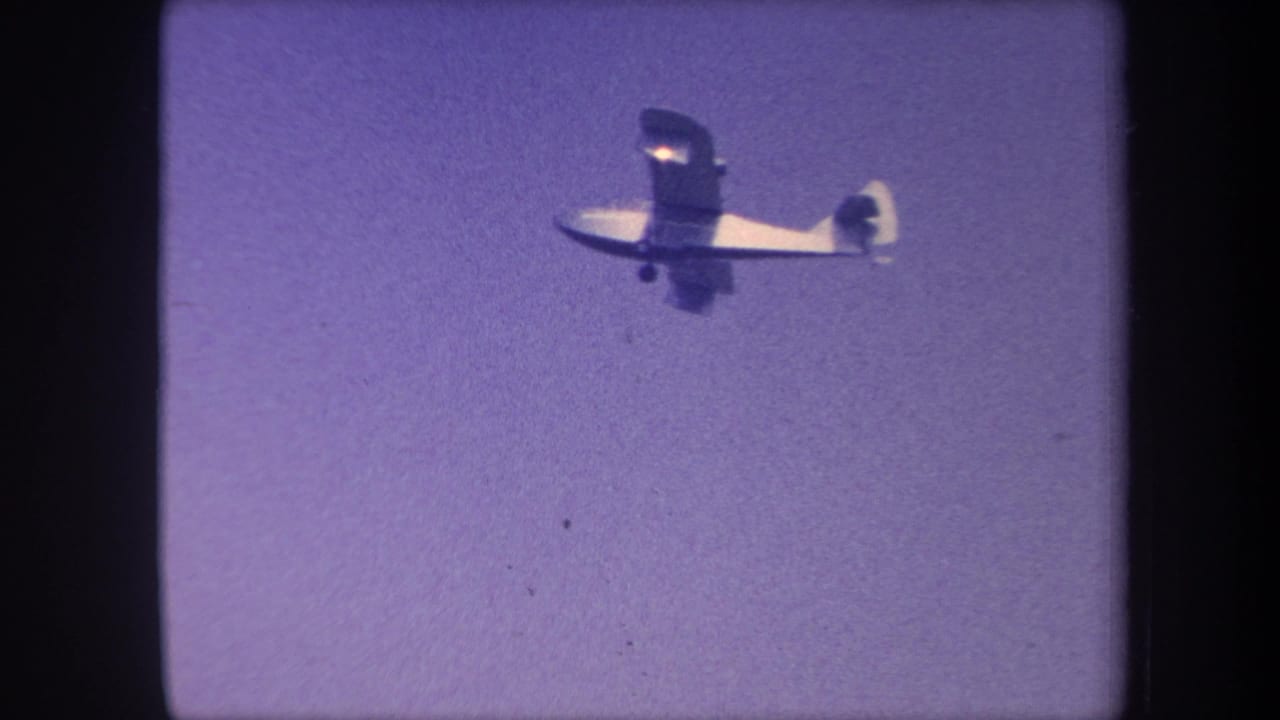 Light single-engine aircraft closer to camera with more fuselage detail visible, against a purple-shifted 8mm sky, circa 1959 California.