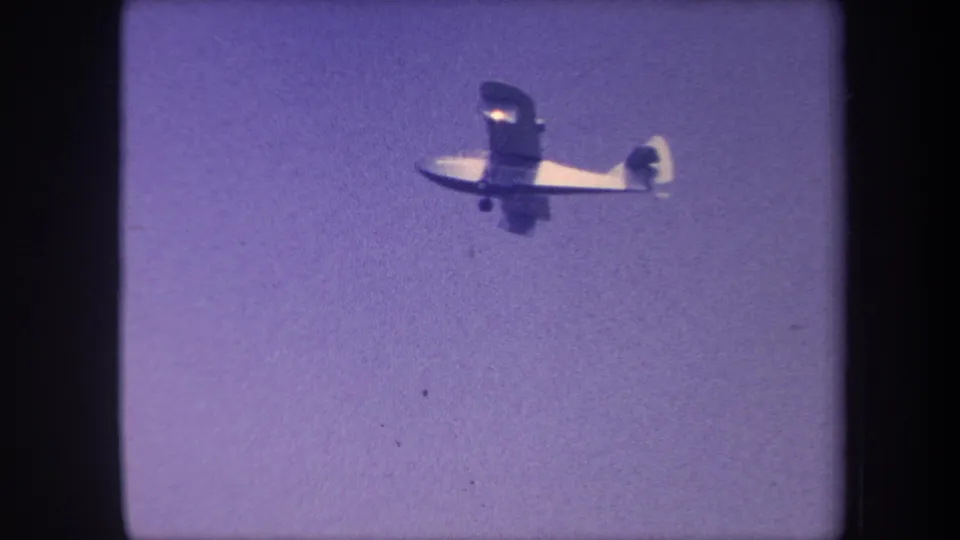 Light single-engine aircraft closer to camera with more fuselage detail visible, against a purple-shifted 8mm sky, circa 1959 California.
