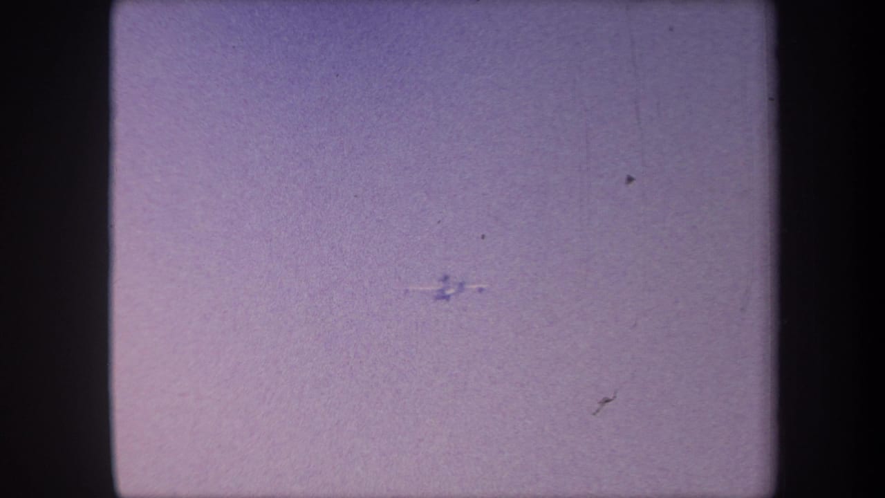 Small aircraft repositioning against a lavender sky in aged 8mm home movie footage, circa 1959, with visible film dust and emulsion scratch.