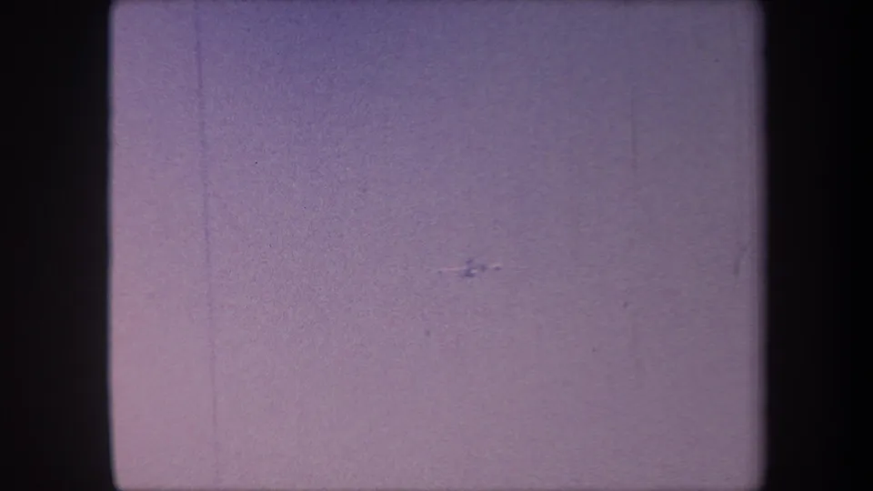 Small aircraft continuing a maneuver against a hazy lavender sky in 8mm home movie footage, circa 1959, with visible emulsion scratches.