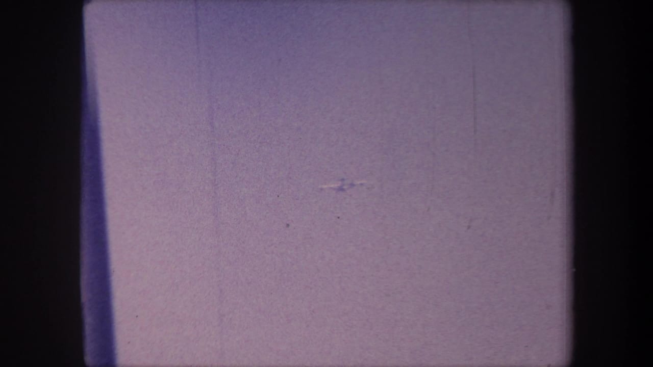 Opening frame of a 1959 8mm home movie; almost entirely consumed by a purple emulsion shift, with a possible faint aircraft speck visible at distance.