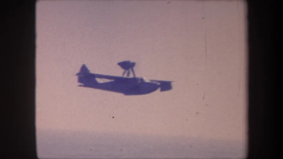 1959 8mm frame from Yucaipa, California: a twin-float seaplane silhouette with a prominent dorsal dome crossing a pale sky from left to right.
