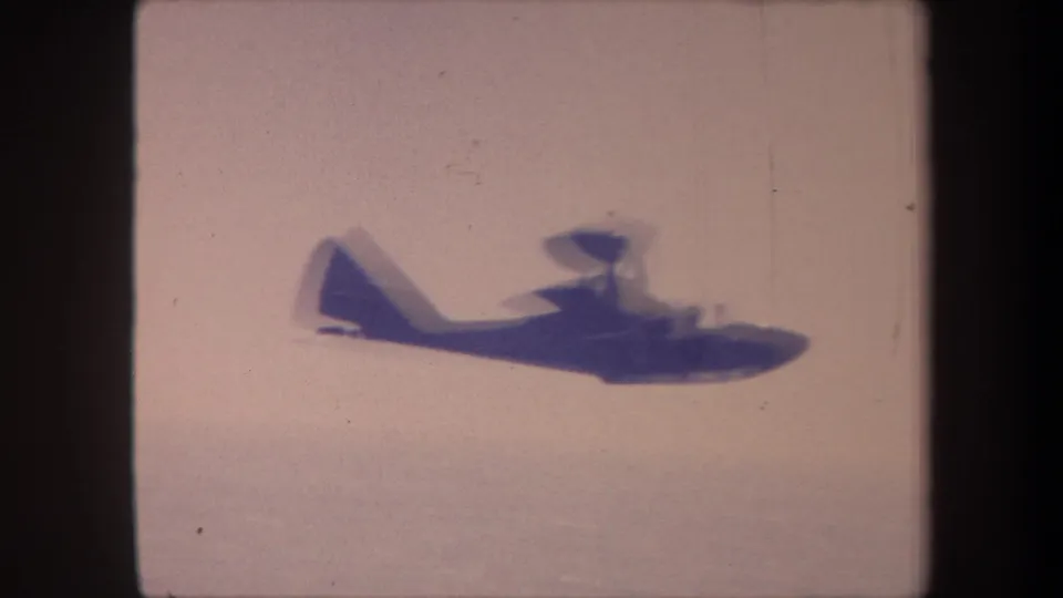 Closing 8mm frame: the seaplane has moved right as the camera pans to follow, with motion blur and coarse grain visible on the silhouette against a pale sky.