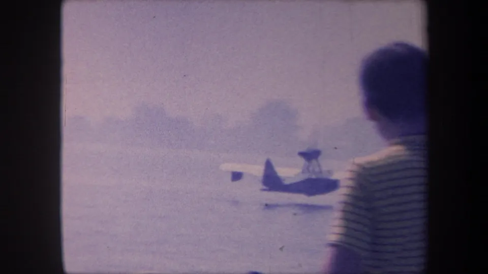 Observer in striped shirt seen from behind watches amphibious plane on water; distant shoreline trees visible, 1959 California 8mm
