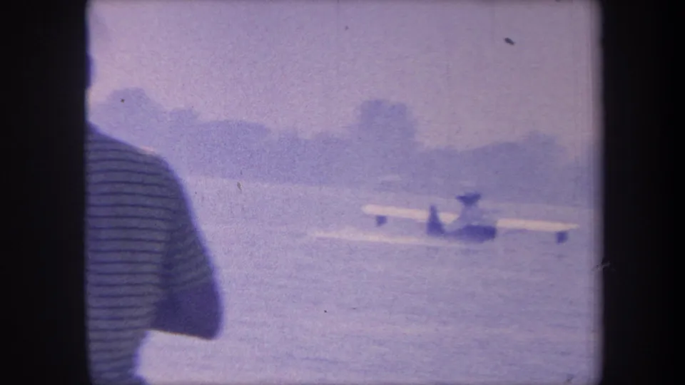 Same observer in striped shirt watches seaplane on water; pontoons clearly on surface, distant treeline visible, 1959 California 8mm
