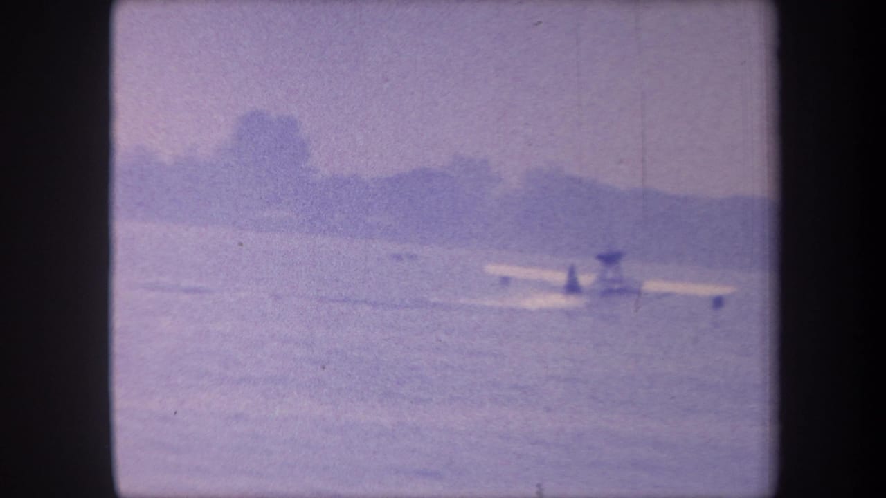 Small amphibious aircraft resting on calm water, distant treeline, purple-cast 8mm frame