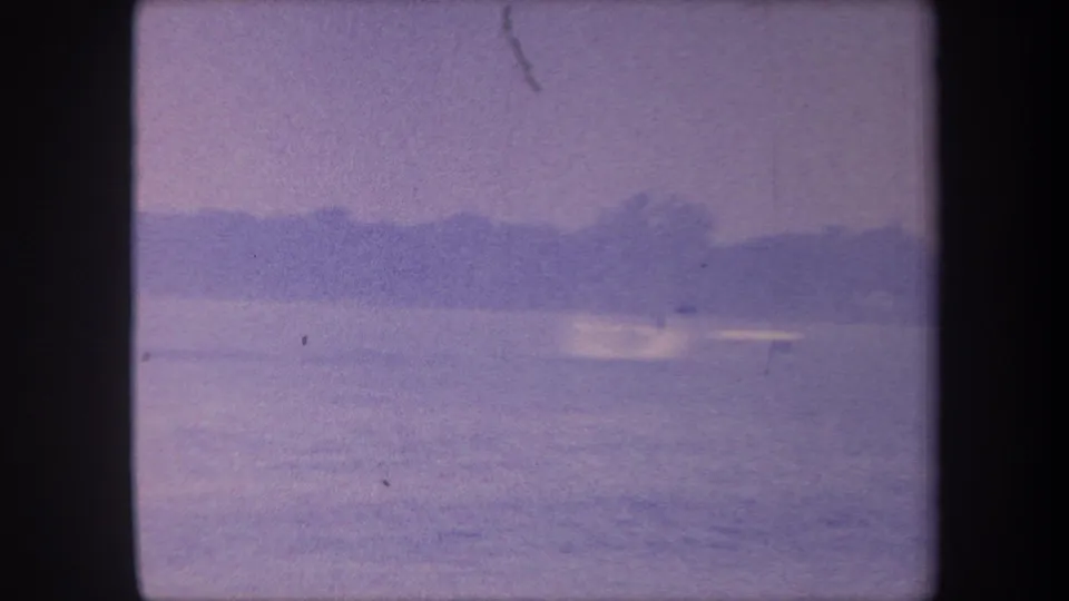 Water surface and low treeline; seaplane appears to have moved off-frame; emulsion scratch visible; late-1950s 8mm color-reversal footage