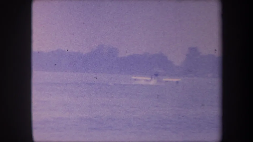 Mostly open water with low treeline; small aircraft or wake faintly visible at distance; late-1950s 8mm color-reversal footage
