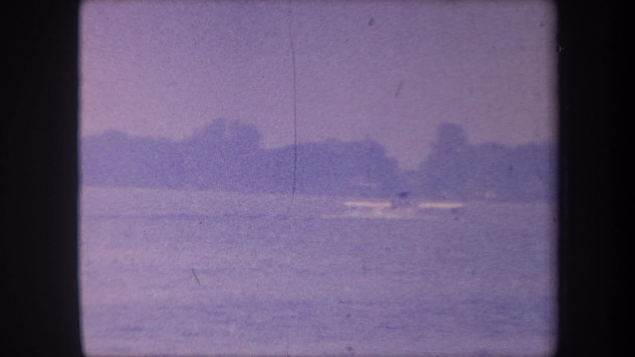 Purple-tinted 8mm frame of a calm lake with a distant treeline and a small white object on the water.