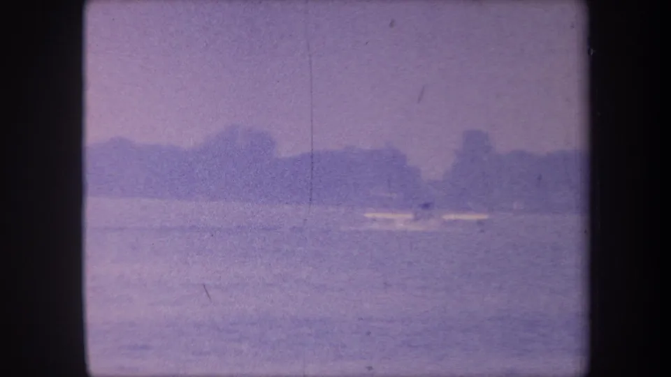 1959 California home-movie frame showing a calm lake, distant treed shoreline, and a small boat on the water, with heavy purple dye-shift and film grain.